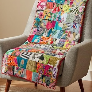 May include: A colorful patchwork quilt draped over a gray armchair. The quilt features a variety of patterned fabric squares in vibrant colors like pink, blue, and green. Red pom-poms trim the edges. The chair has wooden legs.