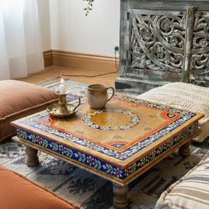 Op de afbeelding: Een lage, vierkante houten tafel met een handgeschilderd ontwerp. De tafel heeft een centraal cirkelvormig patroon en een bloemenrand in blauw, oranje en wit. Een kaars en een mok staan op de tafel, omringd door vloerkussens.