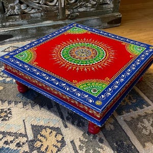 May include: A square, hand-painted wooden footstool. The red top features a central mandala design in green, yellow, and white. The edges are trimmed in blue with white detailing, and it stands on four red, rounded legs.