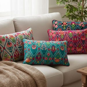 Puede incluir: Un conjunto de cojines decorativos con estampados florales coloridos sobre un sofá beige. Los cojines presentan varios diseños en verde azulado, rosa y rojo, con una manta de color neutro sobre el brazo. Una pequeña mesa de madera está en primer plano.