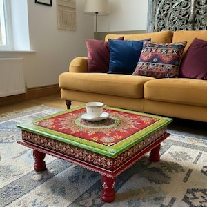 Puede incluir: Una mesa de centro cuadrada con un diseño pintado en rojo, verde y dorado. La mesa tiene patas cortas rojas y un borde decorativo. Una taza de té y un platillo blancos están sobre la mesa. La mesa está sobre una alfombra estampada.