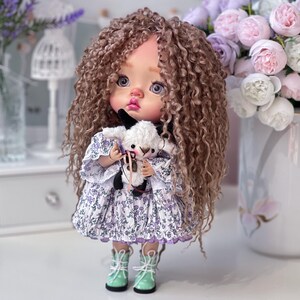 May include: A doll with curly brown hair wearing a purple and white floral dress and green boots. The doll is holding a white stuffed lamb.