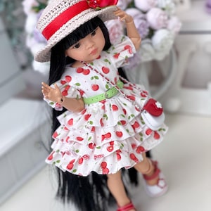 May include: A doll with long black hair wearing a white dress with a red cherry print, featuring tiered ruffles and a light green belt. The doll wears a beige straw hat with a red ribbon and red flower, and white sandals with red accents.