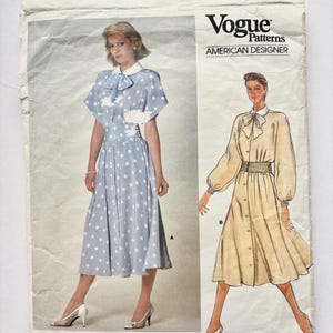 Könnte beinhalten: Vintage Vogue Patterns Schnittmuster mit Designs von Albert Nipon. Der Schnittmusterumschlag zeigt Illustrationen von zwei Kleidern. Eines ist ein hellblaues Polka-Dot-Kleid, das andere ein knöpfbares Kleid. Der Text "Vogue Patterns" und "American Designer" ist sichtbar.