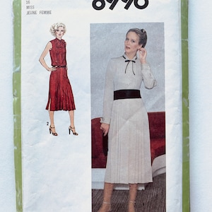 May include: Simplicity sewing pattern number 8996 for a dress. The pattern is for a miss or jeune femme. The pattern shows two models wearing the dress. One model is wearing a red dress with a white collar and a black belt. The other model is wearing a white dress with a black collar and a black belt.