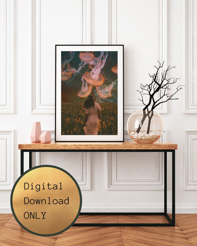 May include: A digital download print of a woman walking through a field of sunflowers with jellyfish floating above her. The background is a dark blue sky with clouds. The image is in a black frame and is on a wooden table.