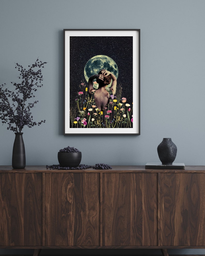 May include: A framed print of a woman standing in a field of flowers under a full moon. The woman is facing away from the viewer and has her arms raised above her head. The moon is a bright white and the sky is dark blue with stars.