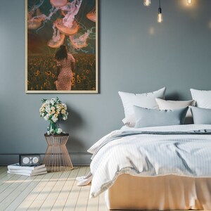 May include: A framed print of a woman walking through a field of sunflowers with jellyfish floating above her. The print is hanging on a gray wall above a bed with white bedding and gray pillows.