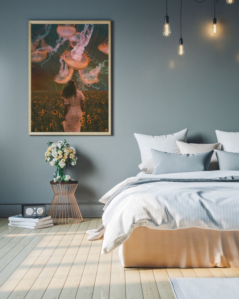 May include: A framed print of a woman walking through a field of sunflowers with jellyfish floating above her. The print is hanging on a gray wall above a bed with white bedding and gray pillows.