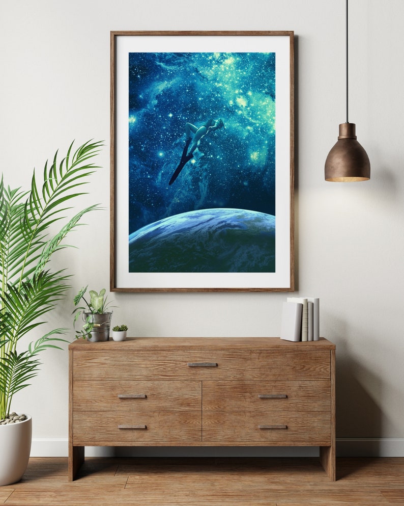 May include: A framed print of a person diving into a blue and green galaxy with stars. The print is hanging on a white wall above a wooden dresser with three drawers.