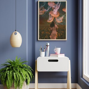 May include: A framed print of a woman walking through a field of sunflowers with jellyfish floating above her. The print is hanging on a blue wall above a white nightstand with a cup of coffee and a thermos on top.