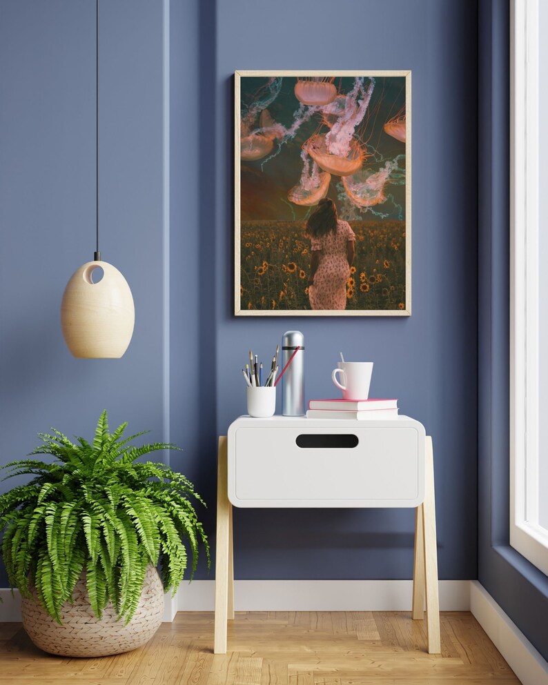 May include: A framed print of a woman walking through a field of sunflowers with jellyfish floating above her. The print is hanging on a blue wall above a white nightstand with a cup of coffee and a thermos on top.