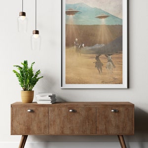Puede incluir: Una impresi&oacute;n enmarcada de un paisaje des&eacute;rtico con tres figuras a caballo que cabalgan hacia una cordillera. Dos platillos voladores est&aacute;n flotando sobre la cordillera, proyectando rayos de luz sobre la escena.