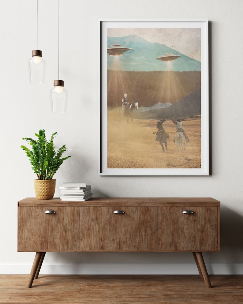 Puede incluir: Una impresi&oacute;n enmarcada de un paisaje des&eacute;rtico con tres figuras a caballo que cabalgan hacia una cordillera. Dos platillos voladores est&aacute;n flotando sobre la cordillera, proyectando rayos de luz sobre la escena.