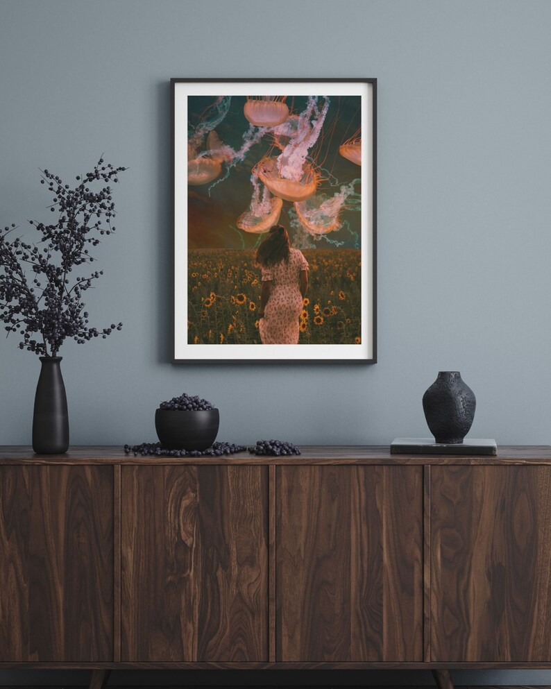 May include: A framed print of a woman walking through a field of sunflowers with jellyfish floating above her. The jellyfish are a pale orange color and the sky is a dark blue with clouds.