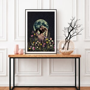 May include: A framed print of a woman standing in a field of flowers with a full moon in the background. The woman is facing away from the viewer and has her arms raised above her head. The moon is a pale yellow color and is surrounded by a dark blue sky with stars.
