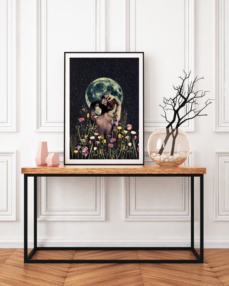 May include: A framed print of a woman standing in a field of flowers with a full moon in the background. The woman is facing away from the viewer and has her arms raised above her head. The moon is a pale yellow color and is surrounded by a dark blue sky with stars.