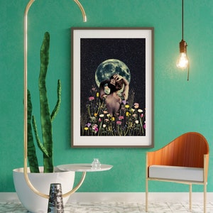 May include: A framed print of a woman standing in a field of flowers with a full moon in the background. The woman is facing away from the viewer and has her arms raised above her head. The print is set against a teal green wall with a cactus plant and a gold metal stand with a white round table top.