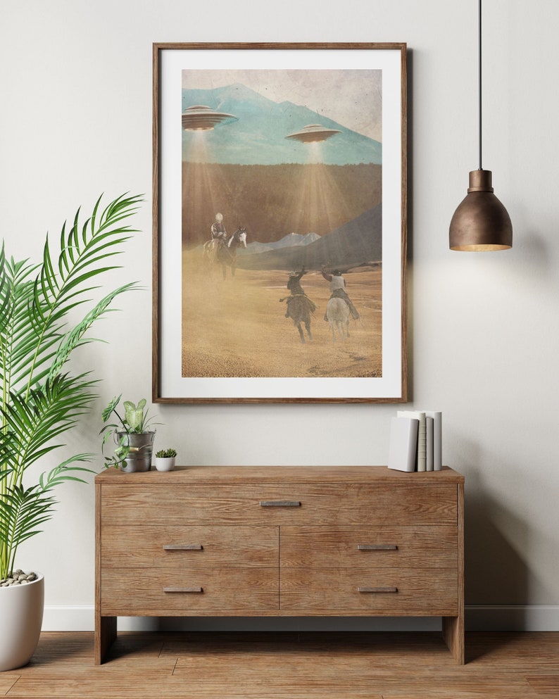 Puede incluir: Una impresi&oacute;n enmarcada de una ilustraci&oacute;n de estilo vintage que representa a vaqueros montando a caballo en un paisaje des&eacute;rtico con tres platillos voladores en el cielo arriba. La impresi&oacute;n tiene un aspecto desgastado y texturizado.