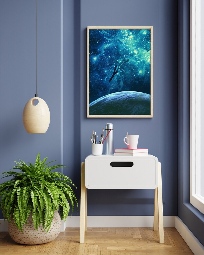 May include: A white nightstand with a drawer and wooden legs. A framed print of a spaceship flying through a galaxy with stars and a planet in the background is hanging on the wall behind the nightstand. A green fern plant is on the floor next to the nightstand.
