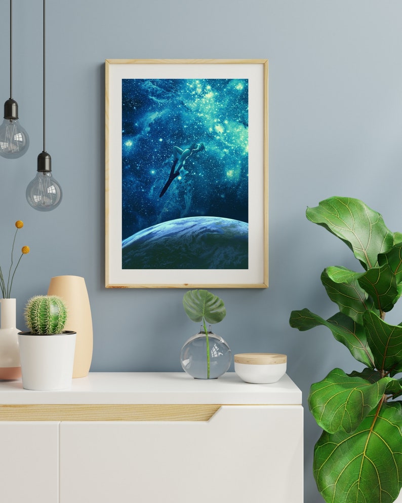 May include: A framed print of a blue and green galaxy with a silhouette of a plane flying over a planet. The frame is light wood and the print is on a white background.