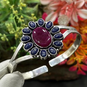Op de afbeelding: Een zilveren armband met een grote, ovale, diep magenta cabochonsteen in het midden. De centrale steen is omringd door een halo van twaalf kleinere, ovale, donkerblauwe lapis lazuli stenen in een bloemachtige opstelling.
