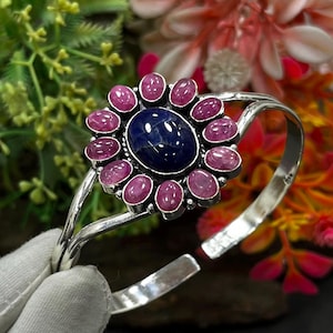 May include: A silver cuff bracelet featuring a large, oval, deep blue central stone surrounded by a halo of twelve smaller, oval, pinkish-red stones. The metal band is polished and open-ended, showcasing a floral-inspired arrangement of gemstones.