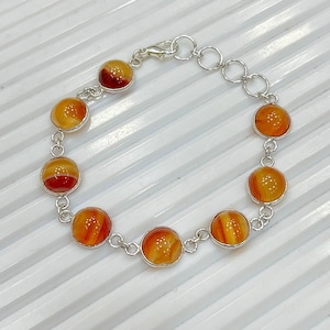 May include: A silver bracelet featuring ten round, polished orange and red agate stones. The stones are set in individual silver bezels and connected by small silver links. The bracelet has a lobster clasp closure.