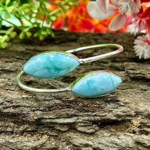 Peut inclure: Un bracelet argenté orné de deux pierres ovales de larimar bleu clair. Les pierres présentent un aspect marbré et sont serties dans un design épuré et ouvert. Le bracelet est présenté sur une surface brune texturée, avec des accents floraux flous en arrière-plan.