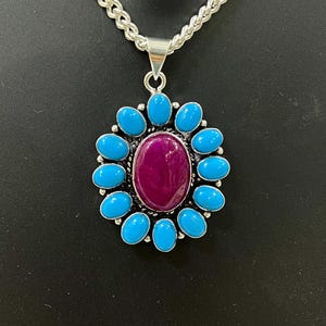 May include: A silver pendant necklace. The pendant has a central oval, deep magenta stone, encircled by turquoise-coloured oval stones. The necklace is suspended from a silver chain.