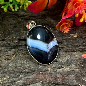 May include: Oval-shaped pendant with a polished black and white agate stone, featuring a band of light blue. The stone is set in a silver-colored metal frame. The pendant is a jewelry item.