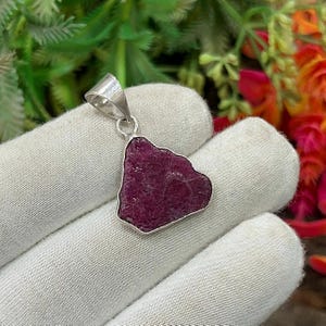 May include: A silver pendant featuring a raw, deep red ruby gemstone. The irregularly shaped ruby is set in a silver bezel and has a silver loop for a chain. The pendant is displayed against a white background.