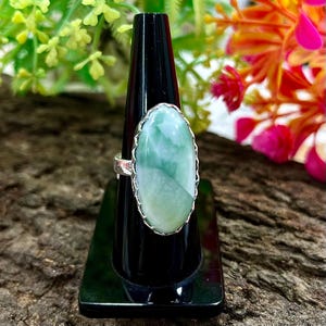 May include: A silver ring featuring a large, oval-shaped, pale green gemstone. The gemstone has a marbled appearance with lighter and darker green areas. The ring is displayed on a black stand against a backdrop of colorful flowers and wood.
