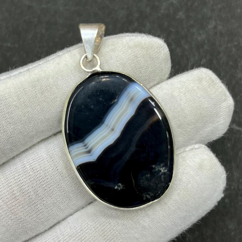 May include: Oval-shaped pendant featuring a polished black agate stone with white and light blue banding. The stone is set in a silver-colored metal bezel and includes a silver bail for a necklace chain.