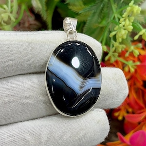 May include: Oval pendant featuring a polished black and white agate stone. The stone has a layered pattern with a band of light blue. The pendant is set in a silver-colored metal with a loop for a chain.
