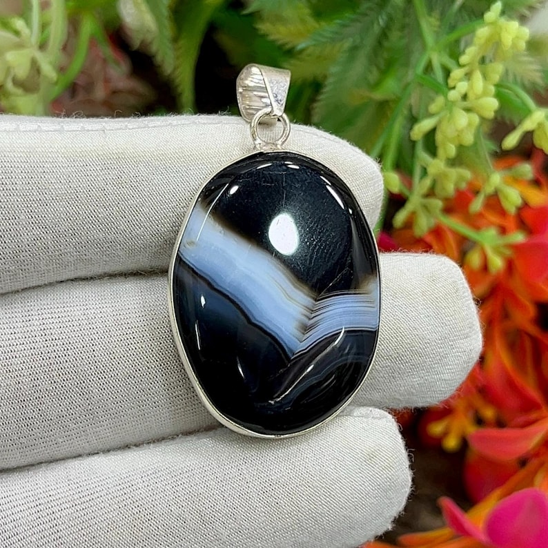 May include: Oval pendant featuring a polished black and white agate stone. The stone has a layered pattern with a band of light blue. The pendant is set in a silver-colored metal with a loop for a chain.