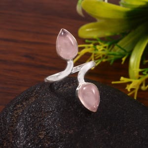 Puede incluir: Un anillo plateado con dos piedras de cuarzo rosa en forma de lágrima. La banda del anillo se curva alrededor de las piedras, creando un diseño único y elegante. El cuarzo rosa tiene un tono rosa suave, y el anillo está sobre un fondo oscuro.