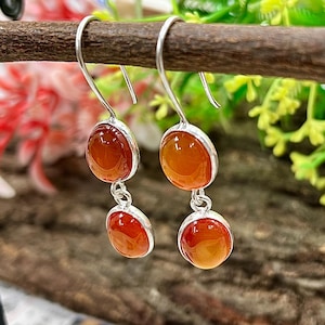 May include: A pair of drop earrings featuring two round, polished, translucent orange carnelian stones set in silver bezels. Each earring consists of a larger top stone and a smaller bottom stone connected by a silver jump ring, hanging from a silver hook.