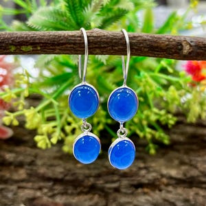 Peut inclure: Une paire de boucles d'oreilles pendantes composées de deux pierres rondes et lisses d'un bleu éclatant serties dans un métal argenté. Chaque boucle présente une pierre plus grande en haut et une plus petite suspendue par un anneau métallique.
