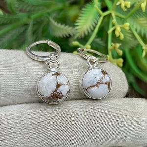 May include: A pair of silver hoop earrings with round, white and brown stone pendants. The earrings are displayed on a light gray fabric with green foliage in the background. The stone has a unique, marbled appearance.