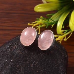 Puede incluir: Un par de pendientes de botón de cuarzo rosa de forma ovalada. Cada pendiente presenta una piedra rosa pálido pulida engastada en un bisel de metal plateado. Los pendientes se exhiben sobre una superficie oscura y texturizada.