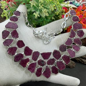 May include: A silver necklace with raw, irregular-shaped ruby stones. The deep red-purple gemstones are set in a silver frame and connected by a silver chain. The necklace is displayed on a white surface.