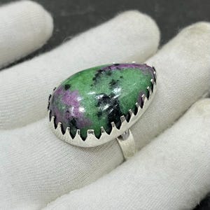 Peut inclure: Une bague en argent ornée d'une grosse pierre ovale. La pierre est un mélange de vert, de violet et de noir, sertie dans une lunette décorative festonnée. L'anneau de la bague est en argent poli simple.