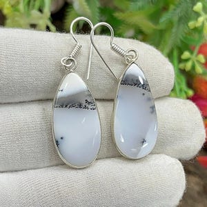 May include: A pair of teardrop earrings. Each earring has a white stone with grey and black dendritic inclusions, set in silver-coloured metal. The earrings have a simple hook design.