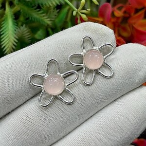 Op de afbeelding: Een paar zilverkleurige oorknopjes in de vorm van vijfbladige bloemen. Elke oorbel heeft een gladde, ronde, lichtroze cabochonsteen in het midden van een draadvormig metalen frame dat de contouren van de bloemblaadjes vormt.