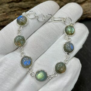 Könnte beinhalten: Ein silbernes Armband mit runden, irisierenden Labradoritsteinen. Die Steine zeigen blaue, grüne und goldene Reflexe. Das Armband ist mit kleinen Silbergliedern verbunden und hat einen Verschluss. Das Armband ist etwa 18 cm lang.