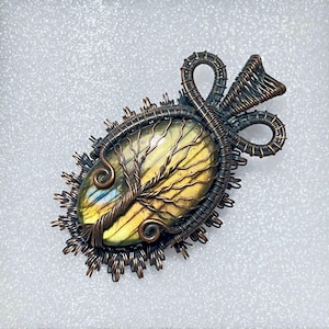 Op de afbeelding: Handgemaakte hanger met een ovale edelsteen in geel en blauw, omwikkeld met koperdraad. Het draadwerk vormt een boomontwerp en een decoratieve balein. Een uniek, door de natuur geïnspireerd sieraad.