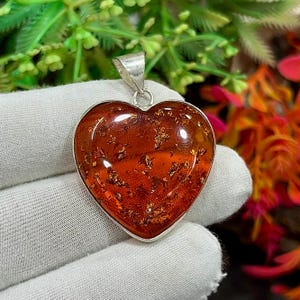 Op de afbeelding: Een hartvormige hanger met een zilveren beugel. De hanger heeft een doorschijnende, rood-oranje steen met gouden vlokken. De steen is gezet in een zilveren rand. De achtergrond is een wit oppervlak met groen en oranje blad.