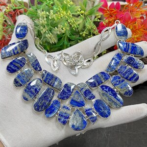 Könnte beinhalten: Eine Statement-Halskette mit mehreren polierten Lapislazuli-Steinen in verschiedenen Oval- und Tropfenformen, die jeweils in einen silberfarbenen Metallrahmen gefasst sind. Die Steine zeigen tiefe Blautöne mit natürlichen grauen und goldenen Einschlüssen.
