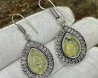 May include: A pair of silver-toned earrings with a teardrop design. Each earring features a light green, oval-shaped stone set within a detailed, openwork frame. The earrings have a hook closure.